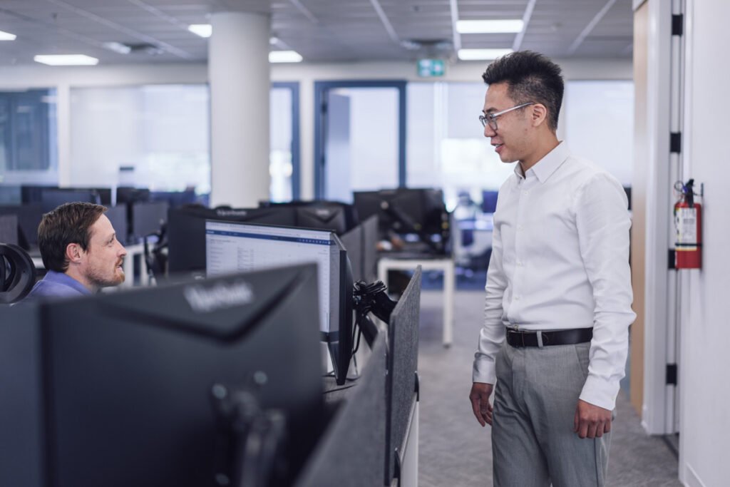 A coworker standing talking to another sitting at their desk